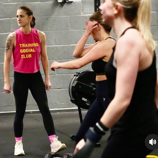 Woman in pink 'Training Social Club' tank top standing next to a stationary bike in a gym setting.